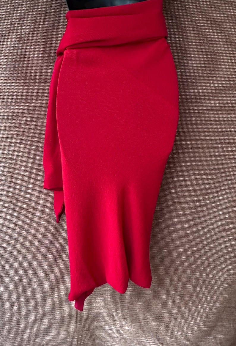 May include: A striking red skirt with a distinctive design. The skirt has a high waist and a draped, flowing fabric, creating an elegant silhouette. The fabric is a smooth, solid colour, suitable for a formal event or special occasion.