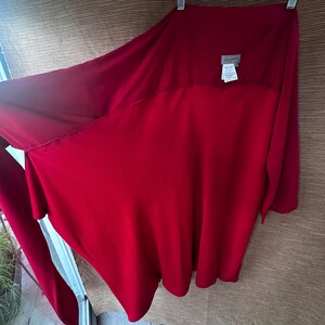 May include: A striking red garment with an unusual, asymmetrical cut. The fabric hangs gracefully, creating a flowing shape. A small label is visible near the neckline. The garment is suspended from a hanger, ready to be worn.