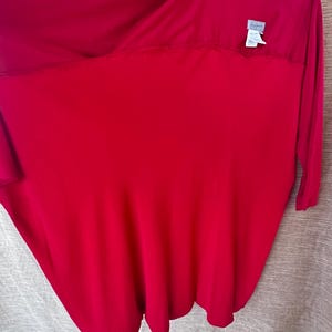 May include: A bright red long-sleeved top with a distinctive, flowing shape. The fabric drapes gracefully, and a small label is visible near the neckline. The top is suspended on a hanger against a neutral backdrop. The garment seems to be made of a soft, lightweight material.