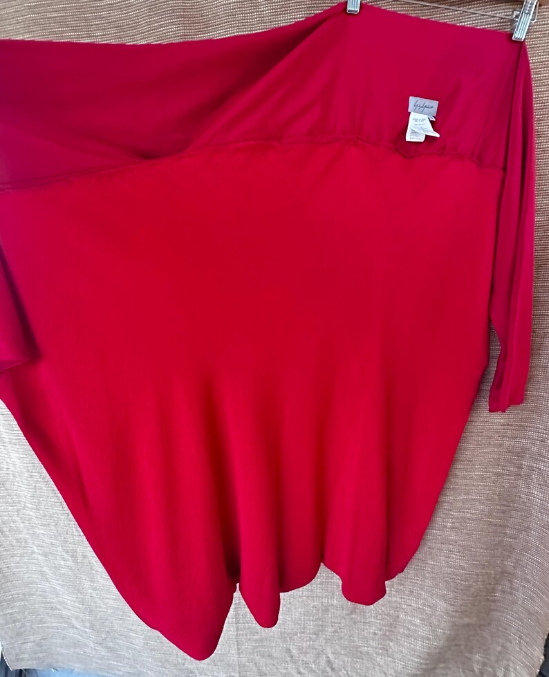 May include: A bright red long-sleeved top with a distinctive, flowing shape. The fabric drapes gracefully, and a small label is visible near the neckline. The top is suspended on a hanger against a neutral backdrop. The garment seems to be made of a soft, lightweight material.