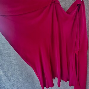 May include: A flowing, magenta-coloured garment, possibly a wrap or skirt, with soft draping and a smooth texture. The fabric is a vibrant, solid colour, and the design suggests a versatile piece of clothing.