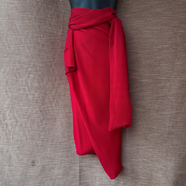 May include: A bright red wrap skirt, displayed on a mannequin. The fabric is smooth and drapes in soft folds, with a long, flowing panel. The skirt is a solid colour, with a simple, elegant design.