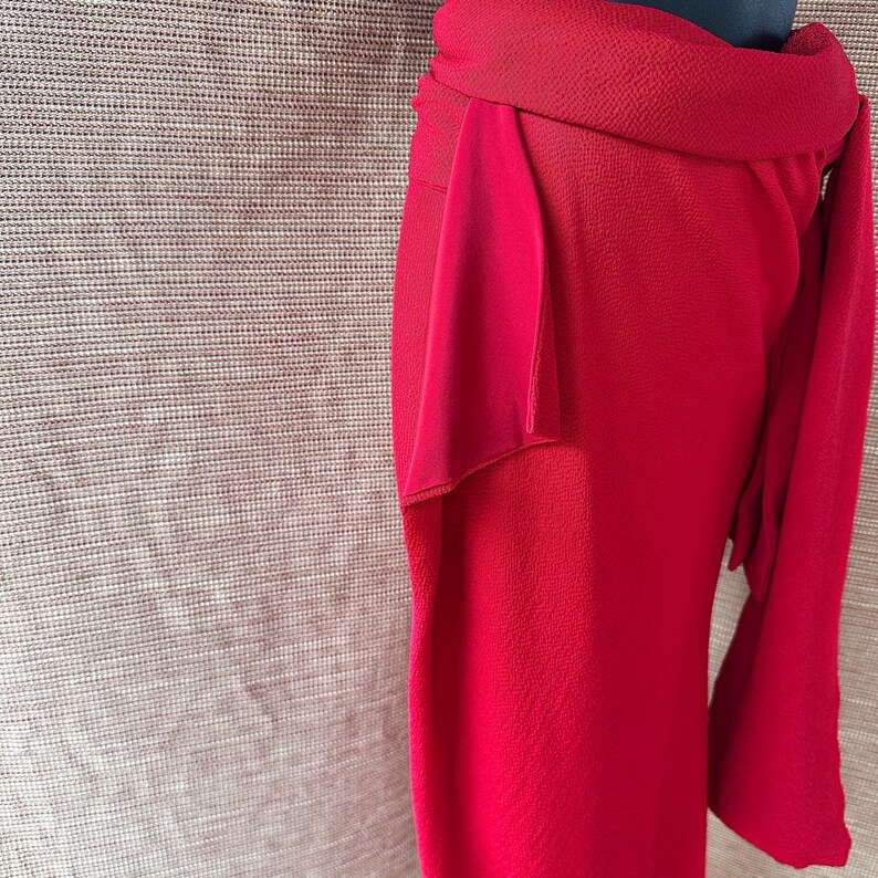 May include: A striking red skirt with an unusual wrap design. The skirt is made from textured fabric and has a flowing, layered detail on the side. It features a wide waistband and a draped element, creating a sophisticated silhouette.