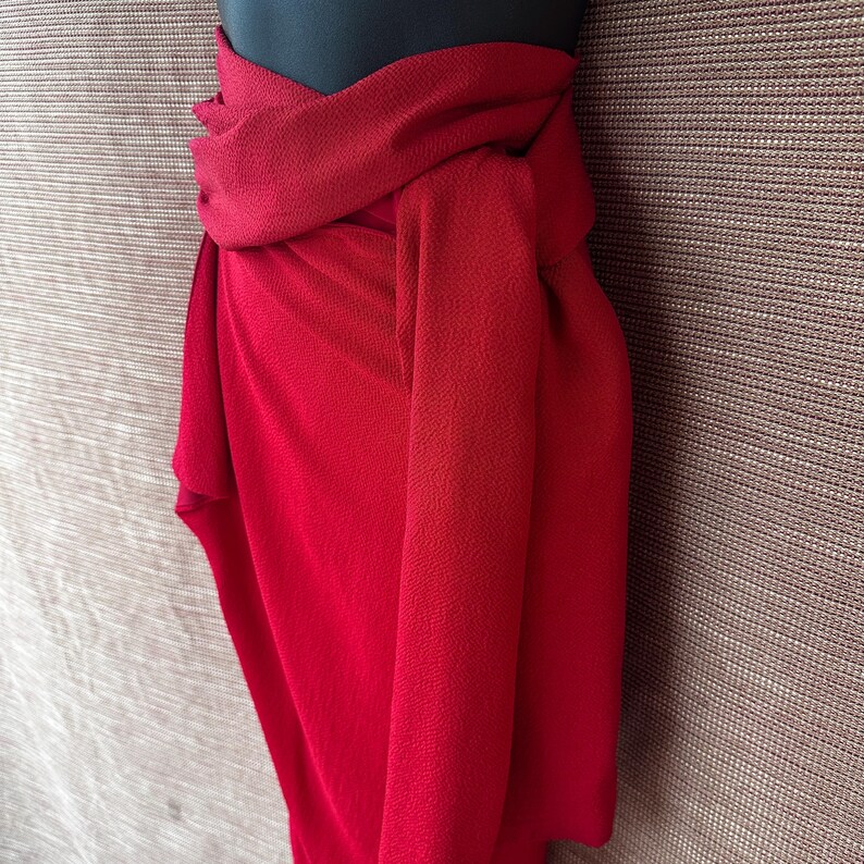 May include: A bright red scarf draped over a black mannequin. The scarf is made of a textured fabric and is knotted at the front. The background is a neutral, textured surface.