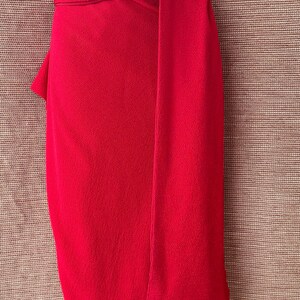 May include: A bright red garment, possibly a skirt or dress, draped over a mannequin. The fabric has a textured appearance, with a wrap-around design and a long, flowing panel. The colour is a bold, solid red.