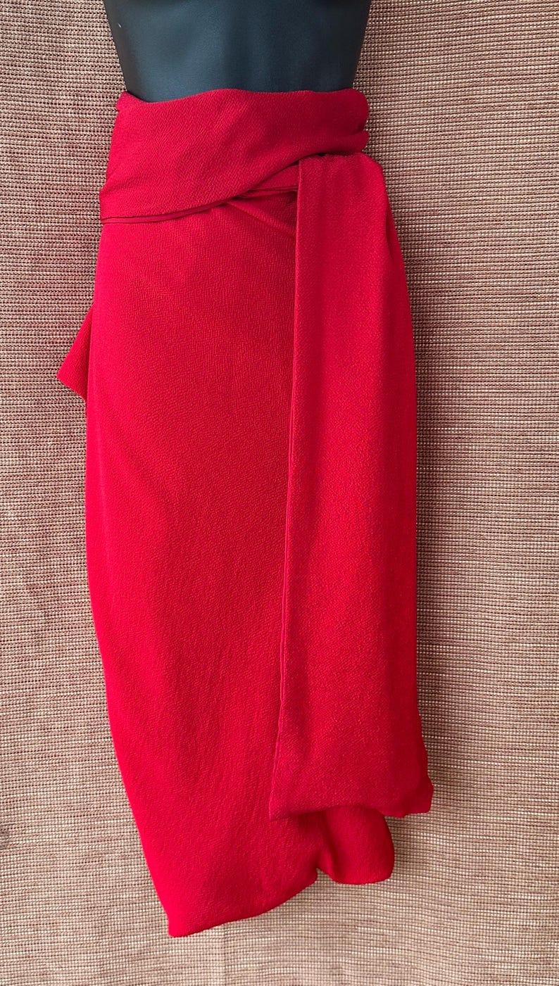 May include: A bright red garment, possibly a skirt or dress, draped over a mannequin. The fabric has a textured appearance, with a wrap-around design and a long, flowing panel. The colour is a bold, solid red.