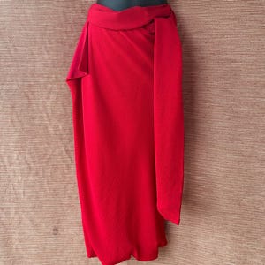 May include: A bright red wrap skirt on a mannequin. The skirt has a high waist and a long, flowing panel that drapes down the front. The fabric has a textured finish. The skirt is a solid colour, without patterns or embellishments. The skirt is a stylish garment.
