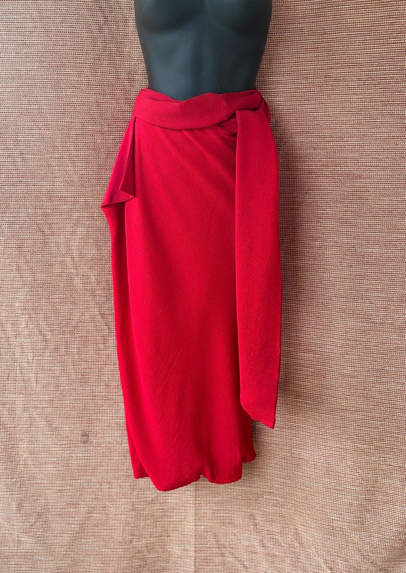 May include: A bright red wrap skirt on a mannequin. The skirt has a high waist and a long, flowing panel that drapes down the front. The fabric has a textured finish. The skirt is a solid colour, without patterns or embellishments. The skirt is a stylish garment.