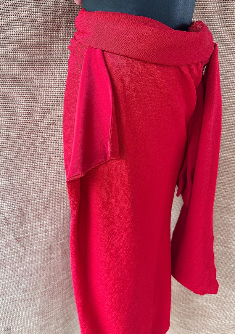 May include: A striking red skirt with a textured fabric and a distinctive wrap design. The skirt has a wide waistband and a flowing, asymmetrical drape, creating a stylish silhouette. The skirt's length is approximately 76 cm.