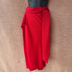 May include: A bright red wrap skirt, knotted at the waist. The skirt is made of a smooth, flowing fabric and is displayed on a mannequin. The design features a long, rectangular panel that hangs down the front.