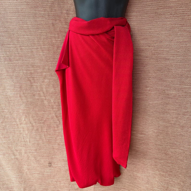 May include: A bright red wrap skirt, knotted at the waist. The skirt is made of a smooth, flowing fabric and is displayed on a mannequin. The design features a long, rectangular panel that hangs down the front.