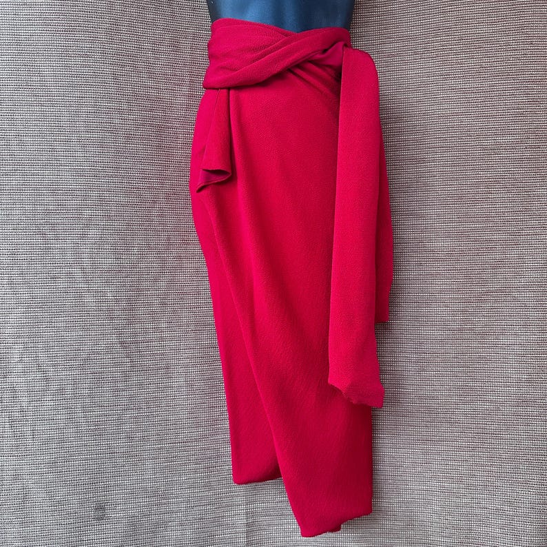 May include: A bright red wrap skirt, tied at the waist with a long sash. The skirt is draped, creating a flowing shape. The fabric is a solid colour, giving a sleek and elegant appearance. The skirt is approximately 90 cm in length.