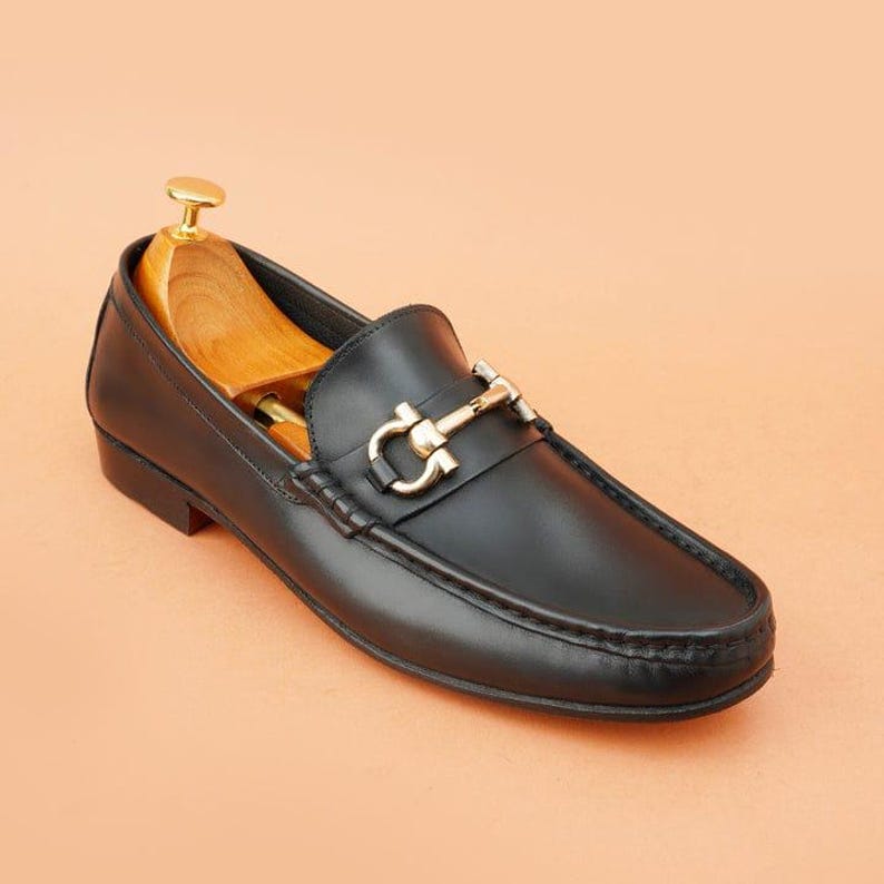K&ouml;nnte beinhalten: Schwarzer Lederloafer mit einer goldfarbenen Metallschnalle. Der Schuh hat eine runde Spitze und einen niedrigen Absatz. Ein h&ouml;lzerner Schuhspanner ist in den Schuh eingesetzt. Der Hintergrund ist einfarbig pfirsichfarben.