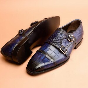 May include: A pair of blue and black leather double monk strap shoes. The shoes feature a textured, crocodile-like pattern and gold-colored buckles. The shoes are displayed on a light peach-colored surface.