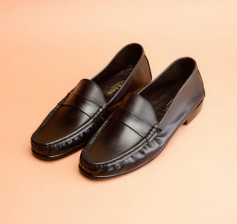 Pu&ograve; includere: Un paio di mocassini in pelle nera dal design classico. Le scarpe hanno una punta arrotondata, una fascia sulla parte superiore e una suola cucita. I mocassini sono esposti su uno sfondo color pesca. Misurano circa 25 cm di lunghezza.