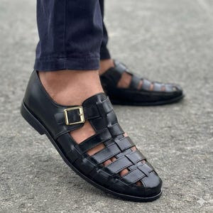 May include: A pair of black leather fisherman sandals featuring a closed-toe woven design, a side strap with a gold-toned metal buckle closure, and a flat sole. The sandals are worn with dark navy blue trousers, showcasing the cutout details.