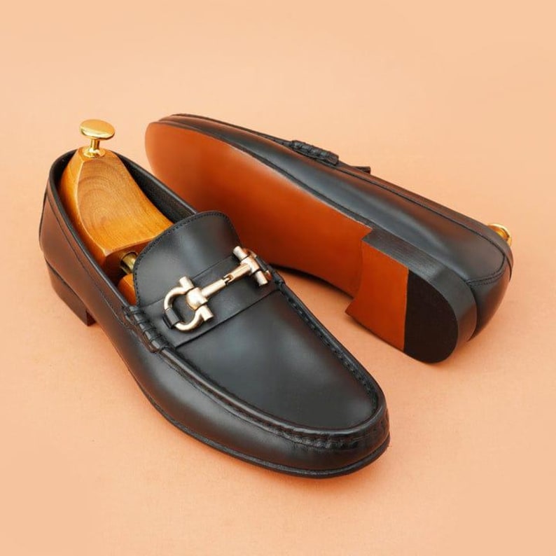 K&ouml;nnte beinhalten: Schwarze Leder-Loafer mit goldfarbener Schnalle. Ein Schuh hat einen h&ouml;lzernen Schuhspanner. Die Sohle ist braun mit schwarzem Absatz. Der andere Schuh liegt auf der Seite und zeigt die Sohle.
