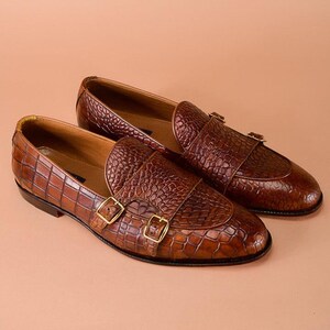 May include: A pair of brown leather loafers with a crocodile texture. The shoes feature double monk straps with gold-colored buckles. The loafers are displayed on a light brown background.