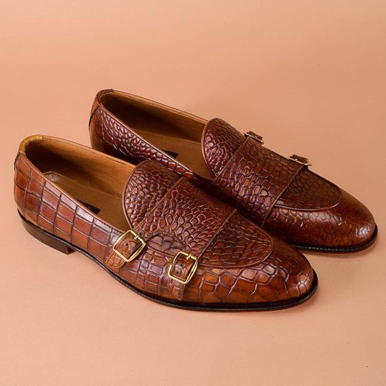 May include: A pair of brown leather loafers with a crocodile texture. The shoes feature double monk straps with gold-colored buckles. The loafers are displayed on a light brown background.