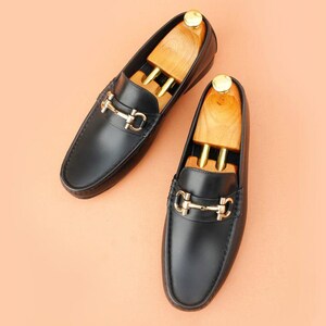 K&ouml;nnte beinhalten: Ein Paar schwarze Leder-Loafer mit goldfarbenen Metallakzenten. Die Schuhe haben ein klassisches Design mit runder Spitze und einem dekorativen Riemen &uuml;ber der Oberseite. Holzschuhspanner befinden sich in den Loafern.