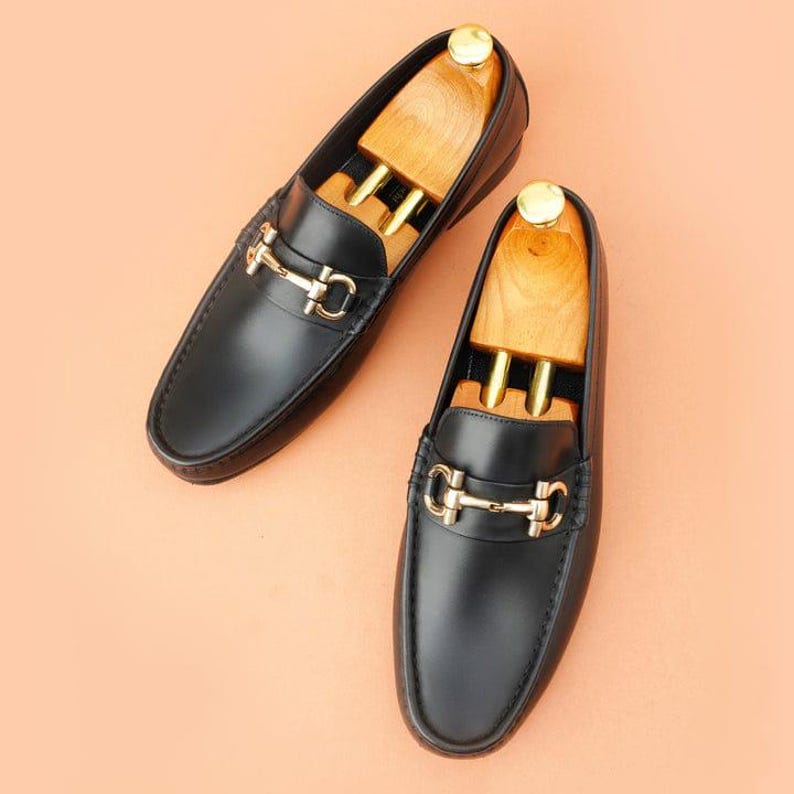 K&ouml;nnte beinhalten: Ein Paar schwarze Leder-Loafer mit goldfarbenen Metallakzenten. Die Schuhe haben ein klassisches Design mit runder Spitze und einem dekorativen Riemen &uuml;ber der Oberseite. Holzschuhspanner befinden sich in den Loafern.
