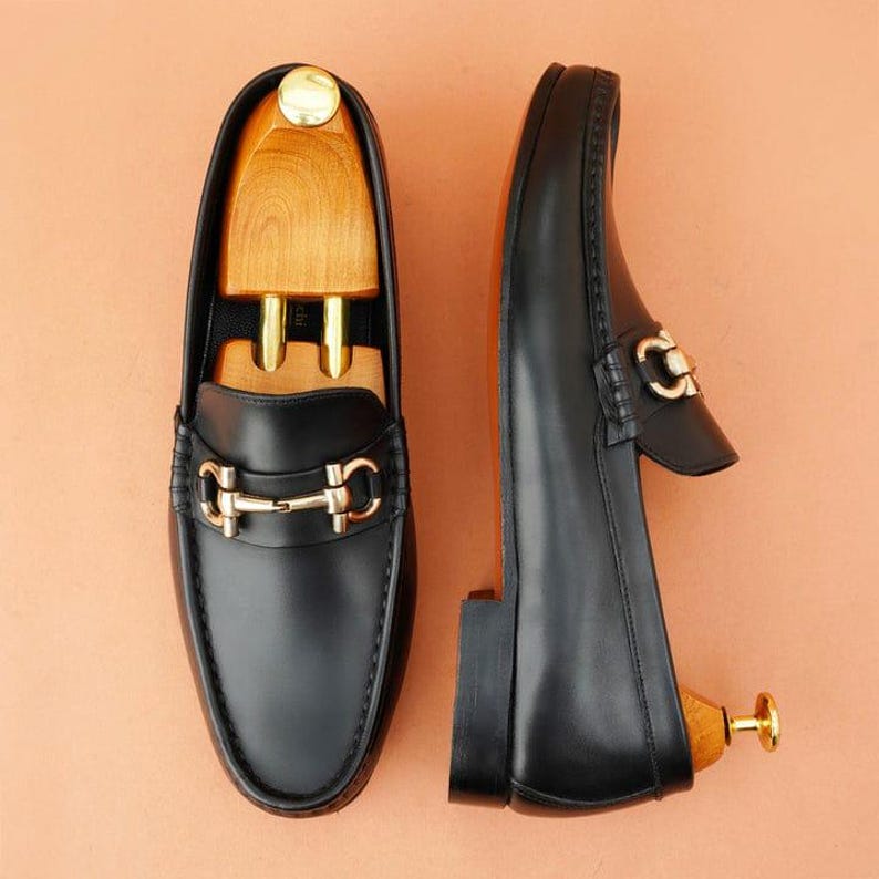 K&ouml;nnte beinhalten: Ein Paar schwarze Leder-Loafer mit goldfarbenen Metallakzenten. Die Schuhe haben ein klassisches Design mit runder Spitze und einem dekorativen Riemen. Schuhspanner aus Holz sind eingesetzt, um die Form der Schuhe zu erhalten.