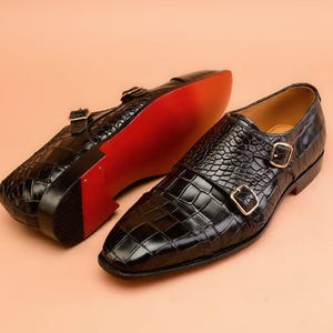 May include: Black leather double monk strap shoes with a crocodile texture. The shoes have gold-colored buckles and a red sole. One shoe is upright, and the other is upside down, showing the sole.
