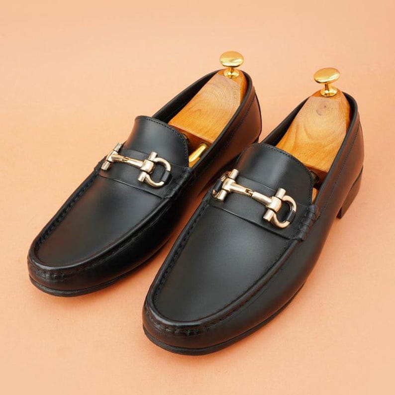 K&ouml;nnte beinhalten: Ein Paar schwarze Leder-Loafer mit goldfarbenen Metallakzenten. Die Schuhe haben ein klassisches Design mit runder Spitze und einem dekorativen Riemen &uuml;ber dem Oberteil. Schuhspanner aus Holz sind in die Schuhe eingesetzt.