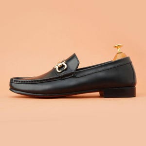 K&ouml;nnte beinhalten: Schwarzer Leder-Loafer mit goldfarbener Schnalle. Der Schuh hat ein klassisches Design mit runder Spitze und niedrigem Absatz. Die Sohle ist rot. Der Schuh wird auf einem h&ouml;lzernen Schuhspanner vor pfirsichfarbenem Hintergrund pr&auml;sentiert.