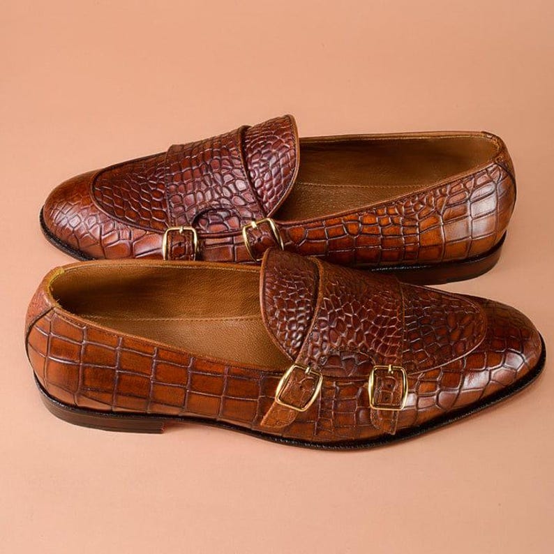 May include: A pair of brown leather loafers with a crocodile skin texture. The shoes feature a double monk strap design with gold-colored buckles. The leather has a glossy finish, and the shoes are set against a neutral background.