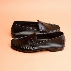 Pu&ograve; includere: Un paio di mocassini in pelle nera. Le scarpe hanno un design classico con punta arrotondata e cinturino sulla tomaia. I mocassini sono presentati su uno sfondo color pesca.
