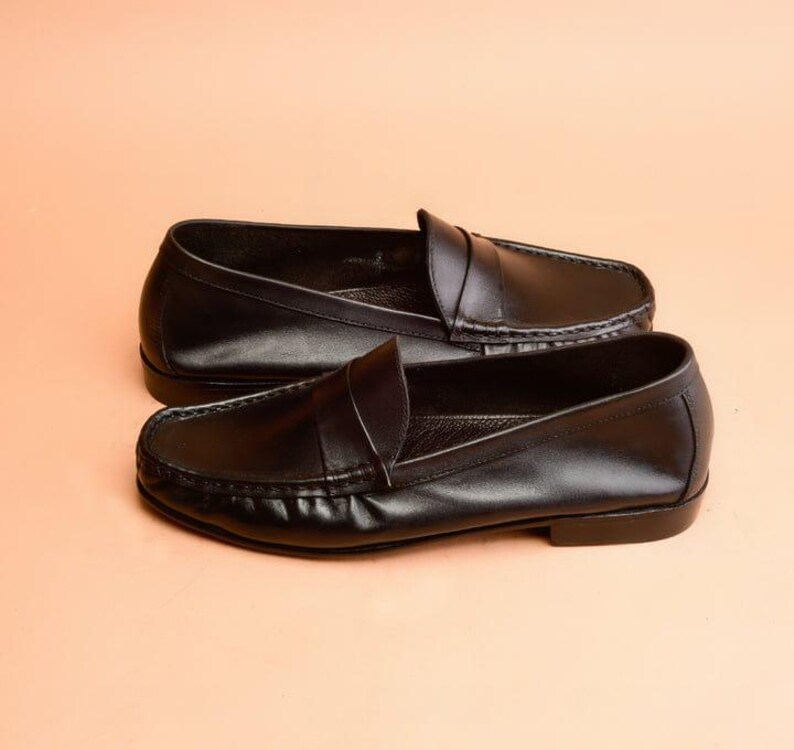 Pu&ograve; includere: Un paio di mocassini in pelle nera. Le scarpe hanno un design classico con punta arrotondata e cinturino sulla tomaia. I mocassini sono presentati su uno sfondo color pesca.