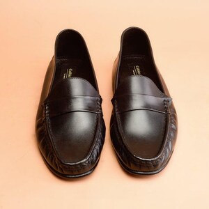 Pu&ograve; includere: Un paio di mocassini neri in pelle. Le scarpe hanno un design classico con punta arrotondata, una fascia sul collo del piede e un tacco basso. Le scarpe sono esposte su una superficie color pesca.