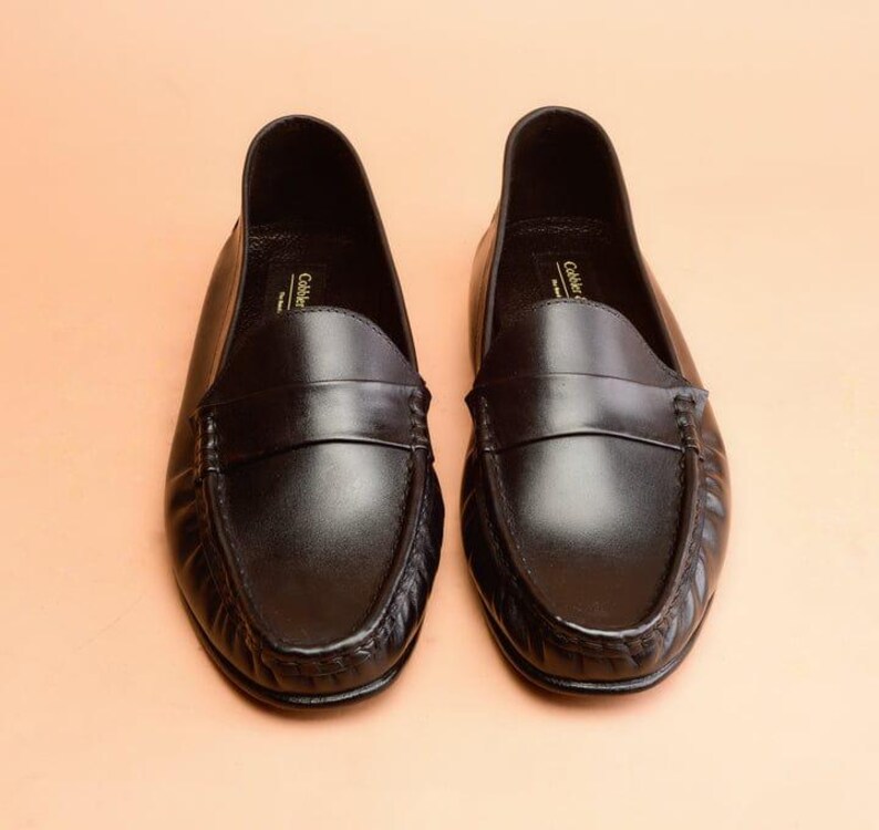 Pu&ograve; includere: Un paio di mocassini neri in pelle. Le scarpe hanno un design classico con punta arrotondata, una fascia sul collo del piede e un tacco basso. Le scarpe sono esposte su una superficie color pesca.