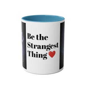 Könnte beinhalten: Weißer Keramikbecher mit blauem Innenraum und Henkel. Der Becher trägt den schwarzen Text "Be the Strangest Thing" mit einem roten Herzsymbol. Der Becher hat auf beiden Seiten einen dunklen Hintergrund.
