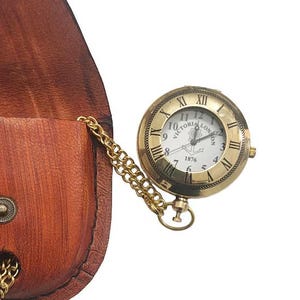 May include: A round brass pocket watch with a white face featuring the text Victoria London 1876 and an anchor symbol, attached to a gold-tone chain. The watch is paired with a brown leather pouch that has a snap button closure and a chain loop.
