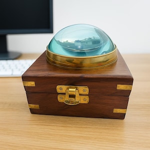 May include: A wooden box with brass detailing, topped with a large, clear magnifying glass. The magnifying glass has a brass rim and a domed shape. The box is secured with a brass clasp.