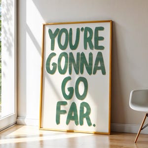 May include: A framed art print with the text "YOU'RE GONNA GO FAR" in green block letters. The artwork has a gold frame and is leaning against a wall. A white chair with wooden legs is to the right.