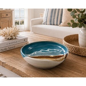 May include: A round ceramic bowl featuring a gradient design that transitions from a deep blue interior with white foam-like patterns to a sandy brown middle section and a solid white exterior base, creating a coastal-inspired aesthetic.