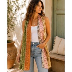 May include: A long, rectangular scarf featuring vertical panels of floral patterns in shades of green, orange, pink, and beige. The scarf is draped over a white tank top and light blue denim jeans, showcasing a multi-colored, botanical print design.