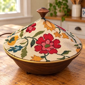 May include: A round, dark wood serving bowl with a conical lid. The lid is covered in a cream-colored fabric featuring a woven floral pattern in red, yellow, and green. A small, dark wooden knob sits at the very top of the conical lid.