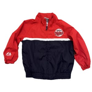 May include: A red and navy blue color-blocked windbreaker jacket featuring a white horizontal stripe across the chest. The Minnesota Twins logo is printed on the upper left chest, and the Majestic brand logo is on the right sleeve cuff.