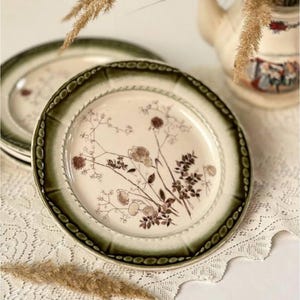 Könnte beinhalten: Ein Satz Vintage-Keramikplatten mit cremefarbenem Mittelteil und dunkelgrünem Rand. Die Teller weisen ein Blumendesign in Brauntönen auf. Die Teller sind auf einer weißen Spitzentischdecke angeordnet.