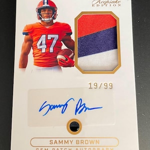May include: A Keepsake Edition sports trading card featuring a photo of a football player in an orange jersey with the number 47, a fabric patch with red, blue, and white stripes, a blue ink autograph, and the text 19/99 and Sammy Brown Gem Patch Autograph.