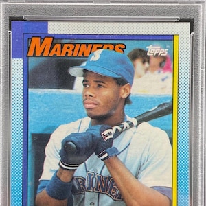 May include: A 1990 Topps Ken Griffey Jr. baseball card, graded NM-MT 8 by PSA. The card features a photograph of the player in a blue Mariners cap and jersey, holding a bat. Text includes 'MARINERS', 'TOPPS', 'KEN GRIFFEY, JR.', and a gold trophy icon.
