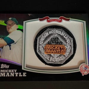 May include: A Topps baseball card featuring Mickey Mantle in a white and green uniform. The card includes an embedded circular patch with the text 1957 AL MVP and MICKEY MANTLE, along with a red banner at the top reading 1957 MVP and a Yankees logo.