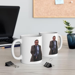 May include: Two white ceramic mugs with a printed image of a man in a suit and tie. The mugs are on a light-colored surface, with a laptop, binder clips, and a small plant in the background.
