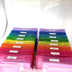 May include: A clear plastic storage case containing two rows of smaller, colorful, translucent plastic boxes. Each box features a white label with black text, including labels such as Word Puzzles, CVC Words, Syllable Sort, and Number Block Addition.