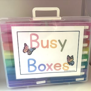 May include: A clear plastic storage case with a white handle, containing colorful boxes. A white sign on the front reads "Busy Boxes" with butterfly illustrations. The boxes are labeled with various educational activity names.