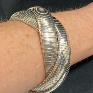 May include: A silver-colored, flexible bracelet with a ribbed design. The bracelet is wrapped around a wrist, showcasing its coiled, snake-like appearance. The metal reflects light, highlighting the texture and form of the jewelry.